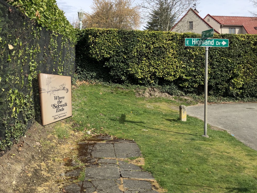 a sidewalk ends and a road turns where a large hedge looms